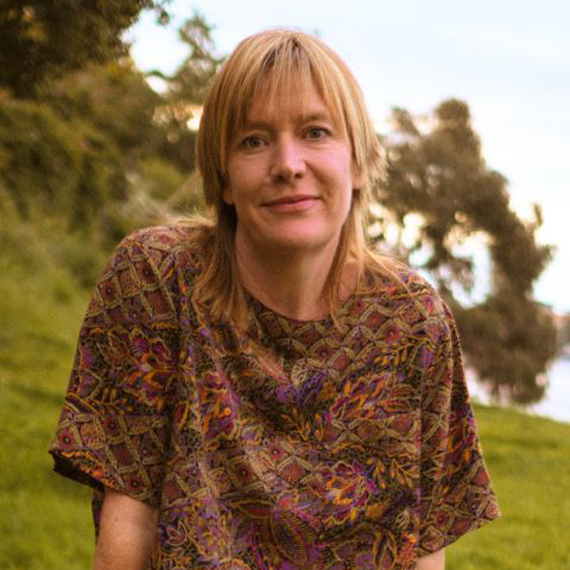 Naturopath Laura Hickey sitting on a large log in a grassy area. She is smiling, has strawberry blonde hair and is in her mid 40's/