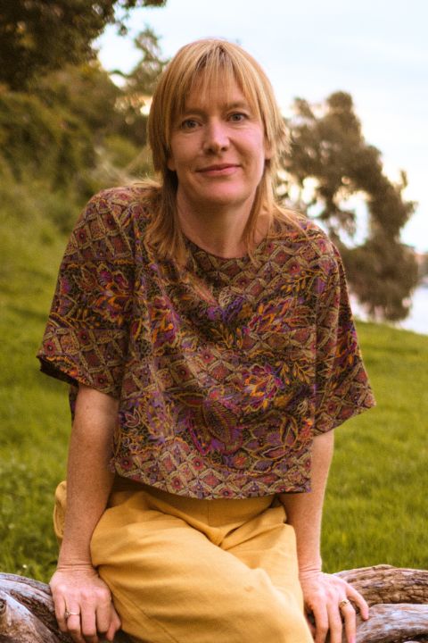 Naturopath Laura Hickey sitting on a large log in a grassy area. She is smiling, has strawberry blonde hair and is in her mid 40's/