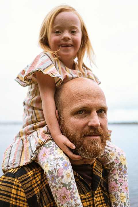 Healthy Young girl with Downs syndrome smiling while sitting on her dads shoulders. Dad has a beard and is also smiling