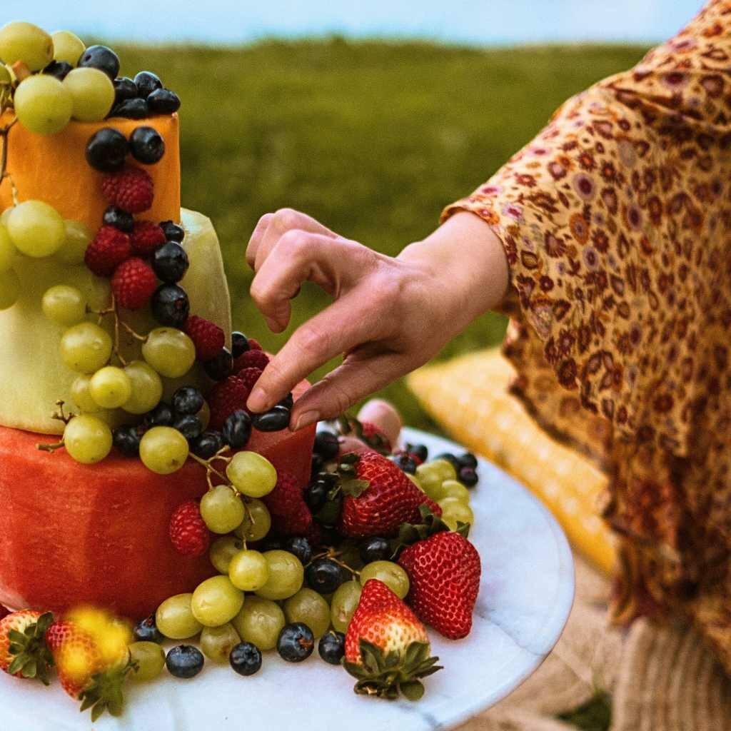 someone is picking a berry from a melon 3 tiered cake, covered in berries and grapes.