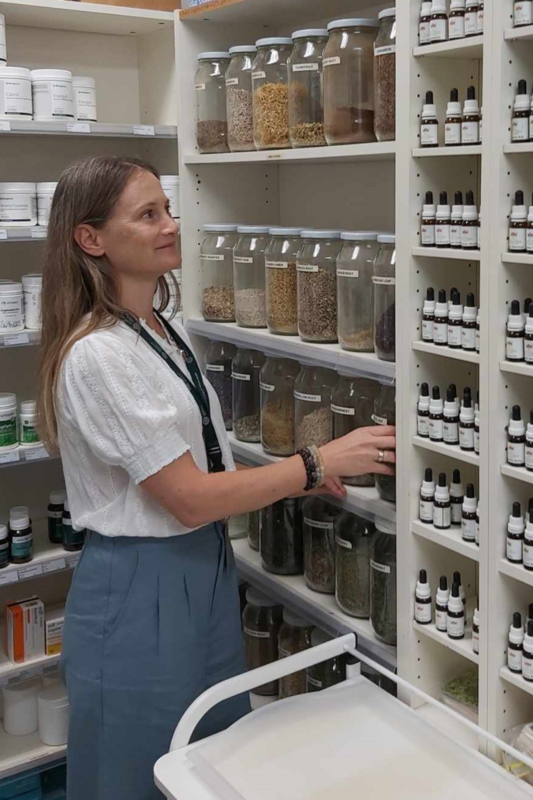 Woman standing in a dispensary with large Jars of dried herbs in front of her, as well bottles of herbs and supplements in containers beside her