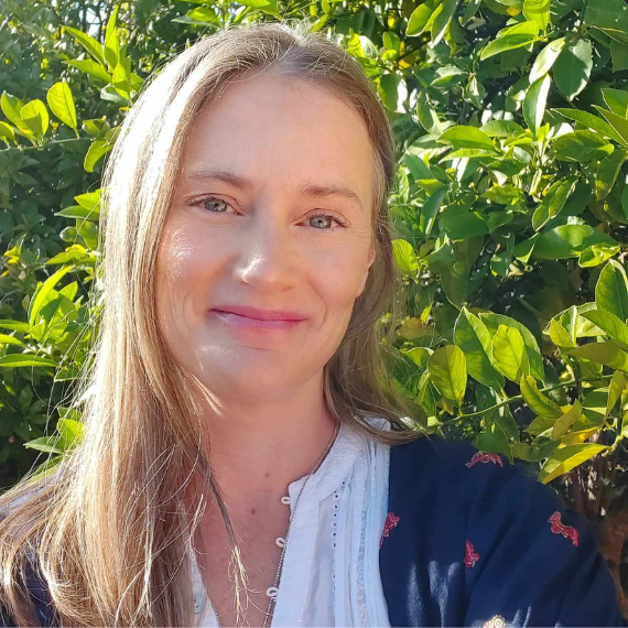 Middle aged woman with long blonde pairing. smiling at the camera, trees and greenery in the background