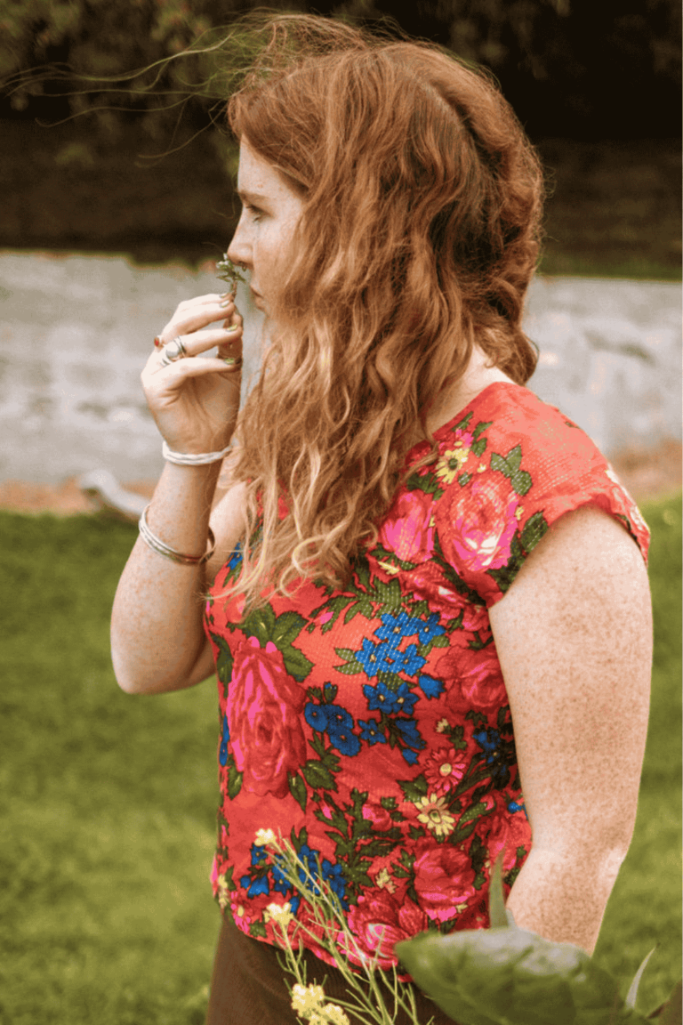 young woman with red hair sniffing a flower. The wind is blowing her hair