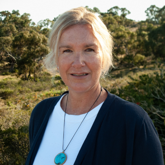 Woman in her 50's with bob length blonde hair. Standing with Australian bush behind her. Wearing a blue pendant necklace