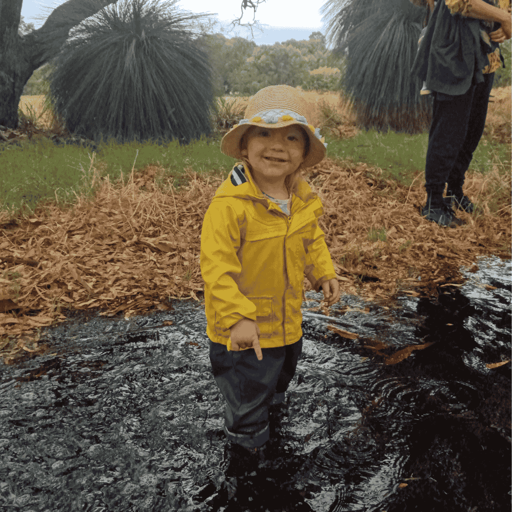 3 year old boyin a big puddle in the bush, with a yellow rain jacket on.