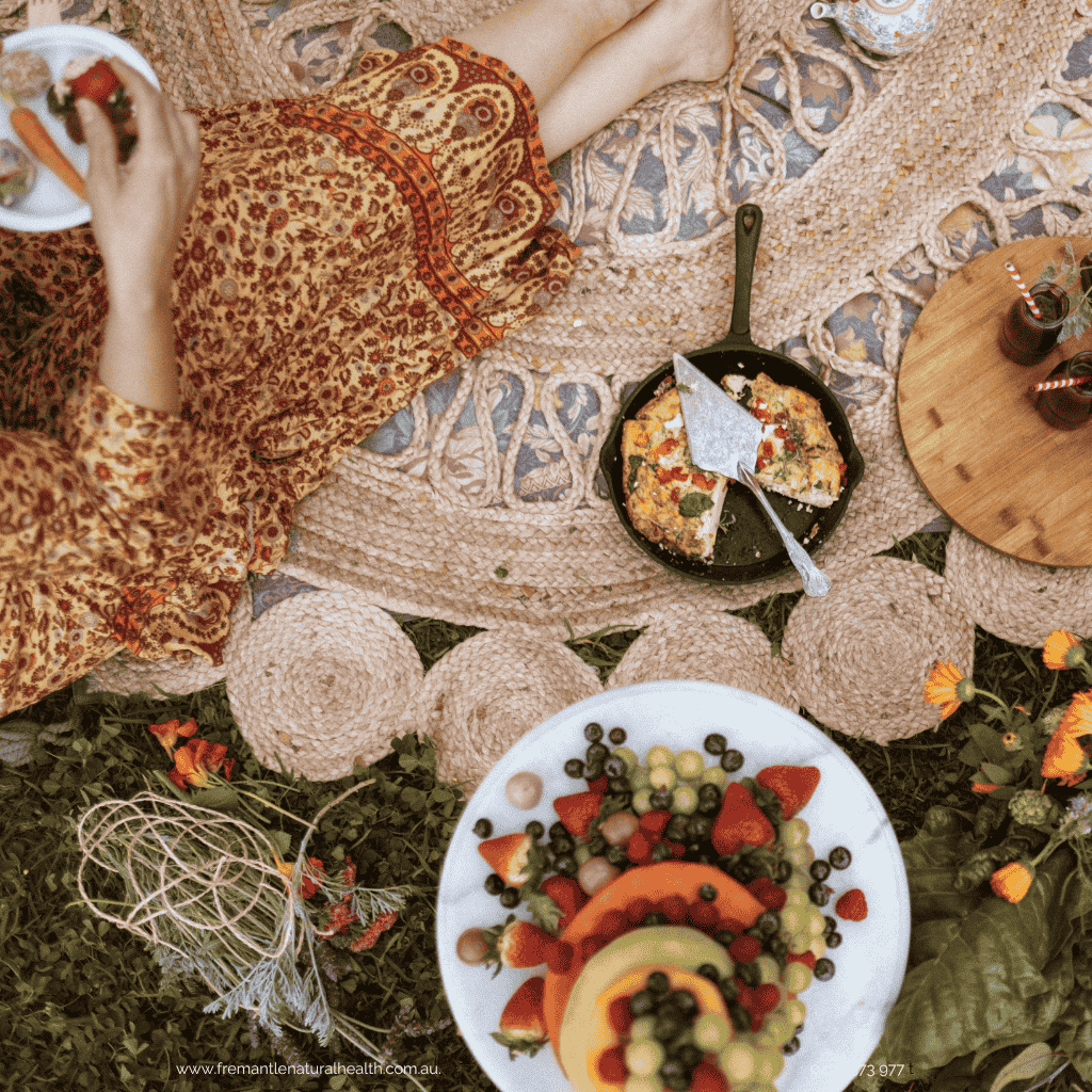 Woman in natural coloured floral dress eating. But you can't see her face. There is a jute round mat with food - pan with a. tortilla and a fruit cake.